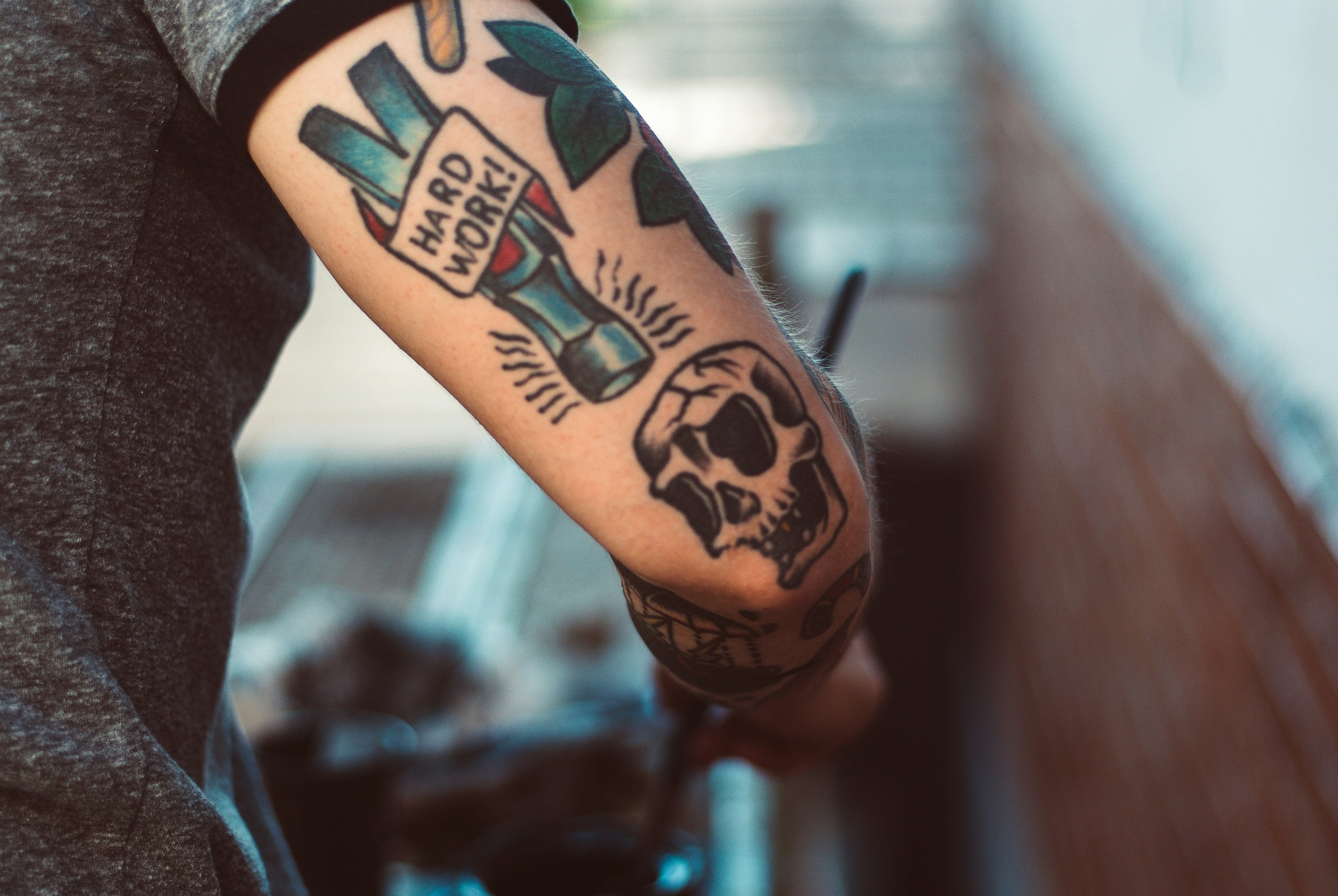 Tattooed arm with various designs including a skull and 'Hard Work' text, blurred background.