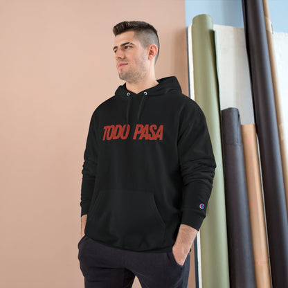 Man wearing a black hoodie with 'TODO PASA' text, standing against a beige wall with rolled-up rugs.