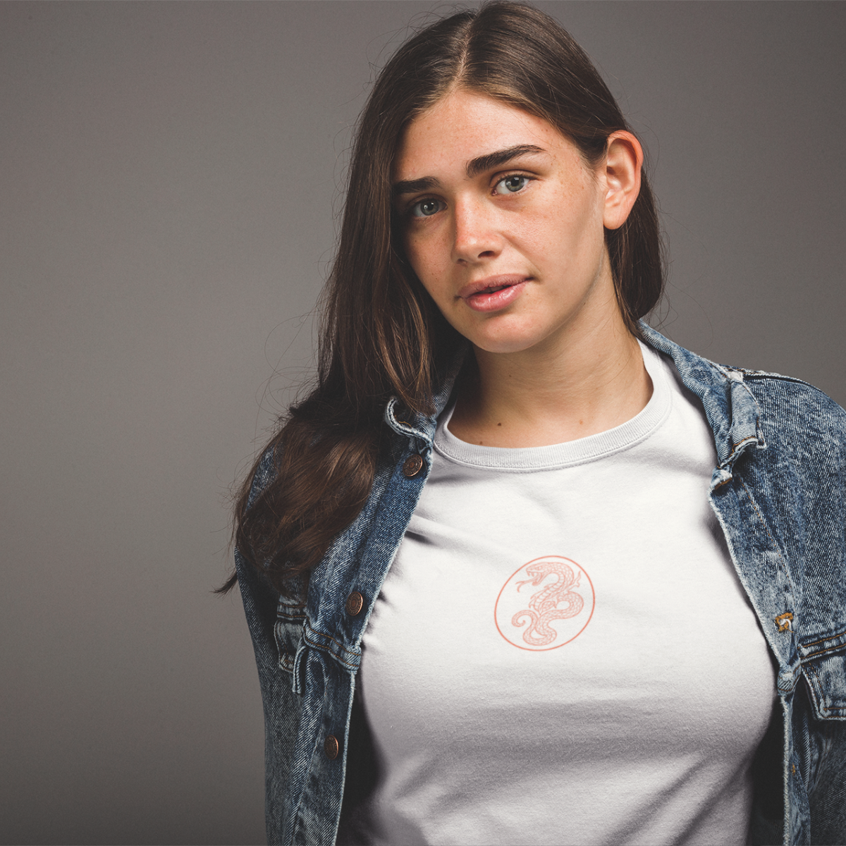 Woman wearing a white t-shirt with a logo and a denim jacket on a gray background