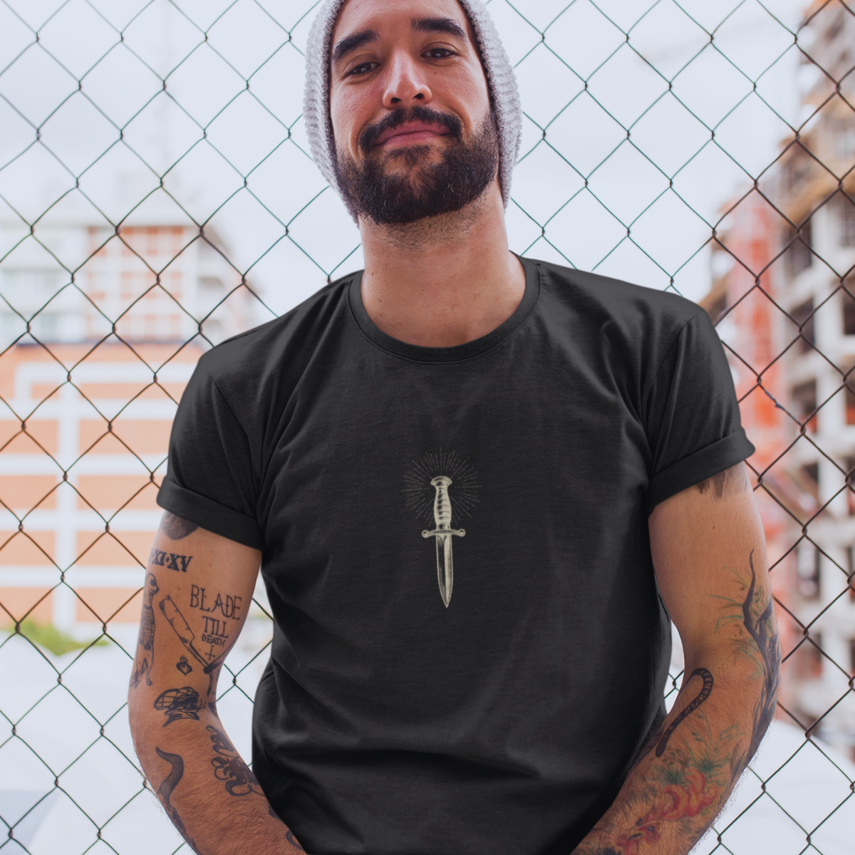 Man wearing a black t-shirt with a dagger design in front of a chain-link fence.